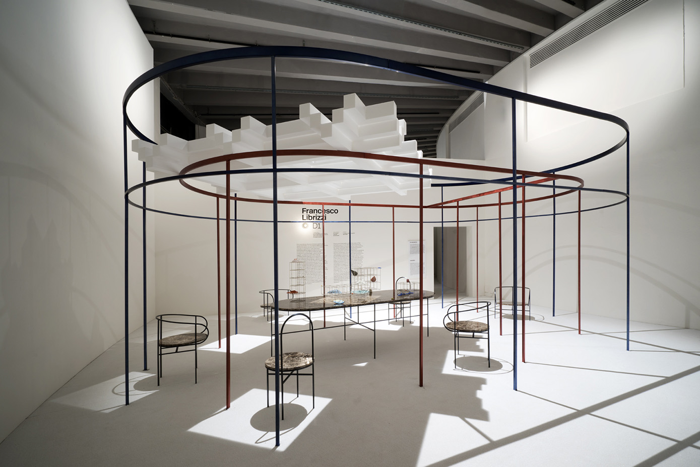 La stanza “D1” di Francesco Librizzi è uno spazio ellittico formato da recinti concentrici fatti di esili colonne di metallo colorato, un modo di raccontare la scoperta dello spazio domestico e il ruolo dell’architettura di mediazione tra paesaggio, spazio domestico e oggetti.